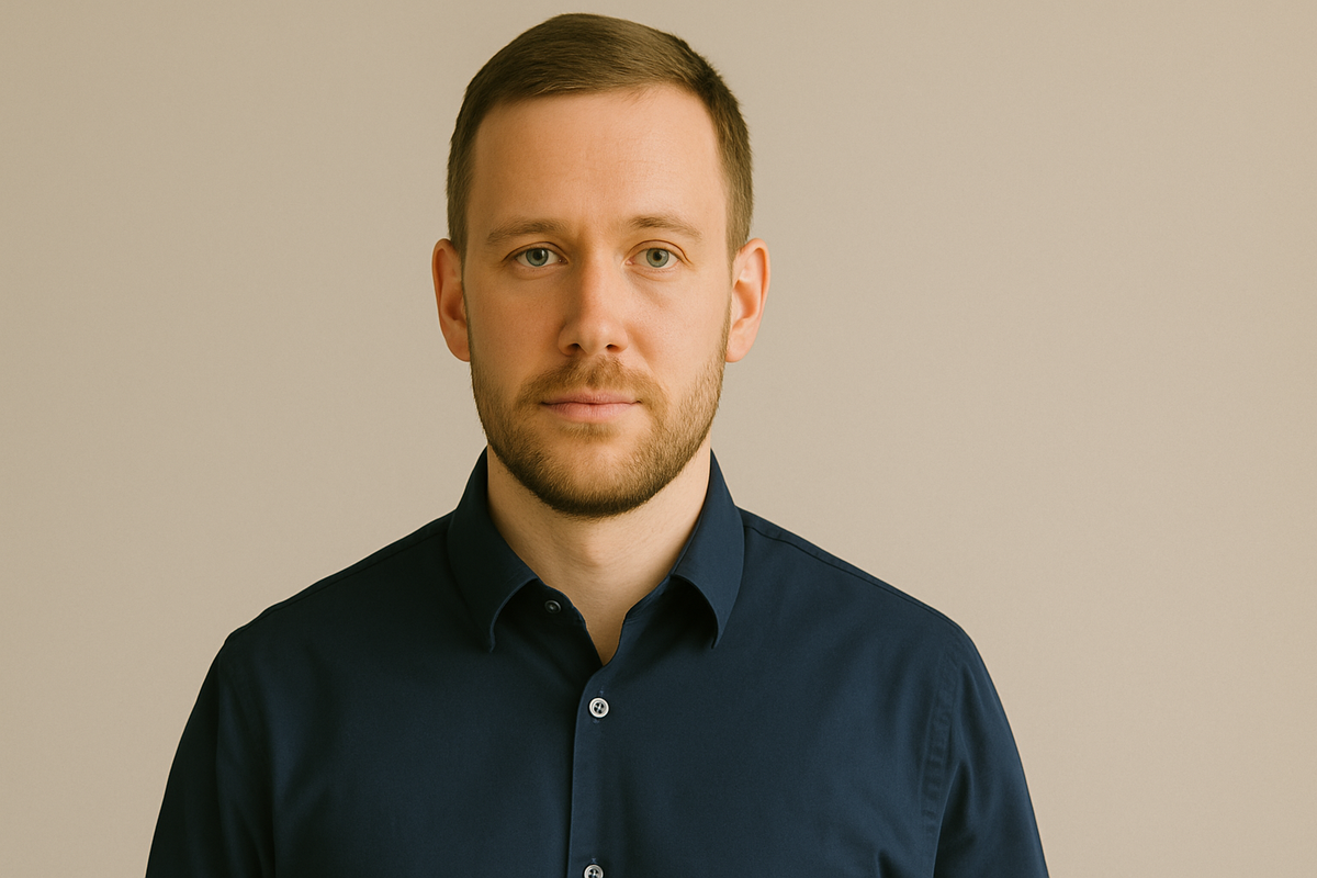 Professional headshot of man in navy shirt in soft daylight, plain background, mid-tone clothing.