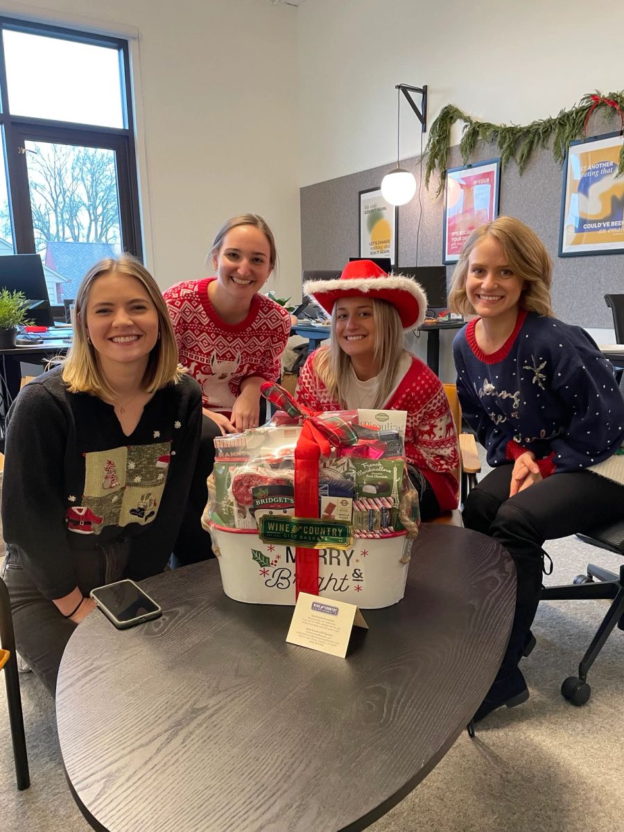 Four Sproutbox teammates in holiday sweaters smiling around a big holiday gift basket in the office