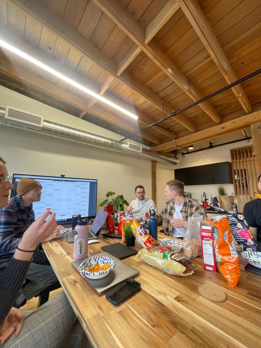 Sproutbox team taste-testing chips and snacks around the office table, with a salty snack bracket on the TV