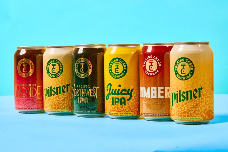 Six colorful Talking Cedar Brewing beer cans lined up on a teal backdrop