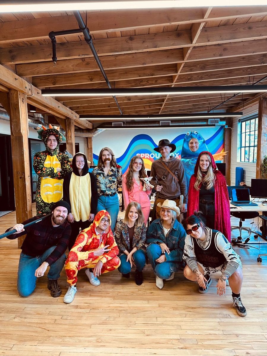 Sproutbox team in Halloween costumes, including a dragon, a fish, and a pizza onesie, in front of the Sproutbox mural