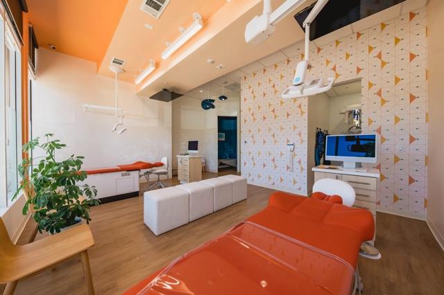 Poppy Kid Dental open bay featuring an orange dental chair in the foreground with overhead lights, dental equipment on the right, a desk with a computer, and a comfortable seating area with a couch and wooden chair. The walls are decorated with a geometric pattern and a large window provides natural light.