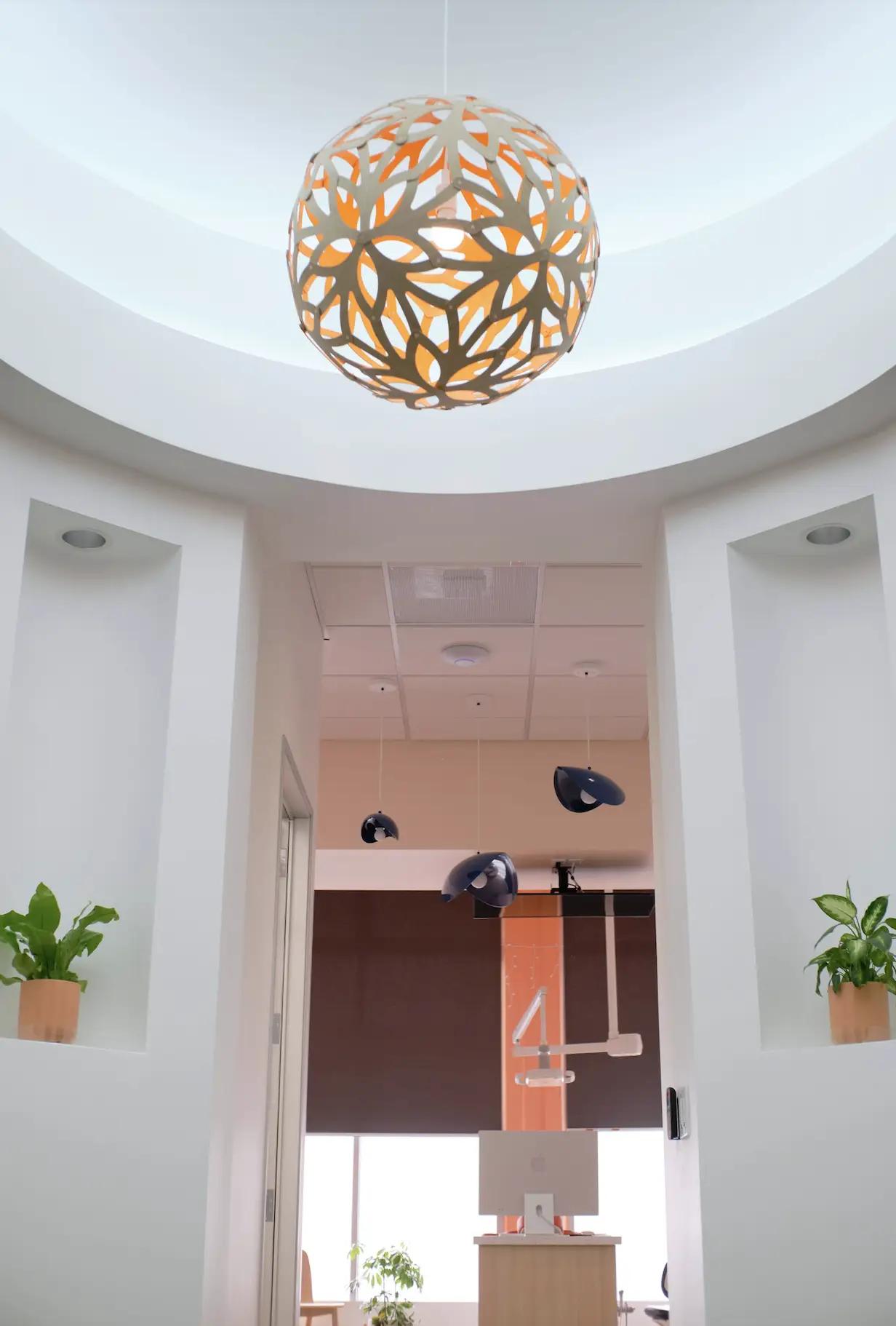 The corridor and rotunda in Poppy Kids Dental with light walls and wooden flooring leading to a brightly lit reception area. An intricate orange spherical light fixture hangs from the ceiling, adding a warm, decorative touch. Natural light streams in from the front windows, and there are small green plants on wooden pedestals, creating a welcoming and serene environment.