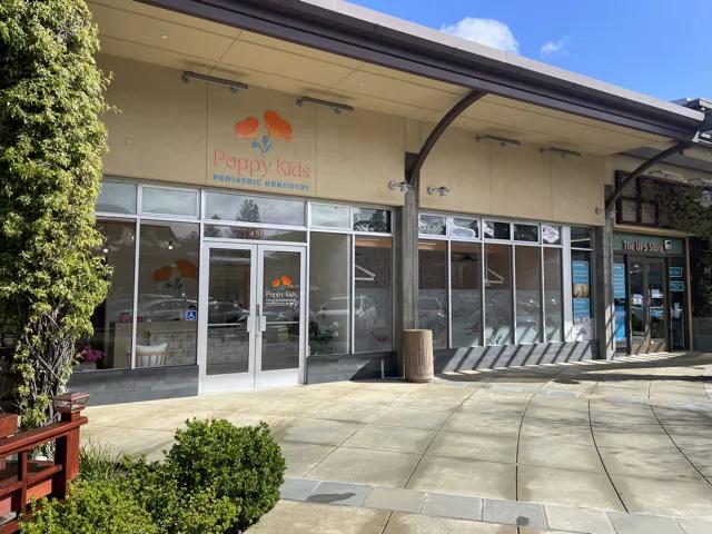 Exterior view of the Poppy Kids Pediatric Dentistry office. The facade is a beige building with a large sign above the entrance featuring the 'Poppy Kids' logo with two orange poppy flowers. The glass doors have the same logo and lead into a lobby.