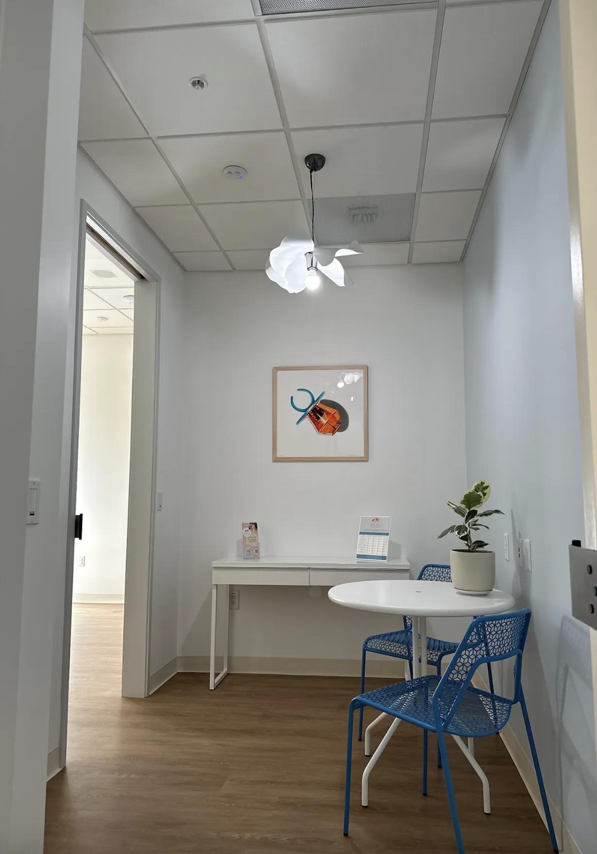 Poppy Kids dental consultation room with pale blue walls and wooden flooring. A white modern desk with information leaflets and a round table with two blue patterned chairs are placed in the room. A potted plant sits on the table, adding a touch of greenery. On the wall, there's a framed artwork of a colorful pacifier made out of candy in orange color, and above, a unique white floral pendant light fixture illuminates the space.