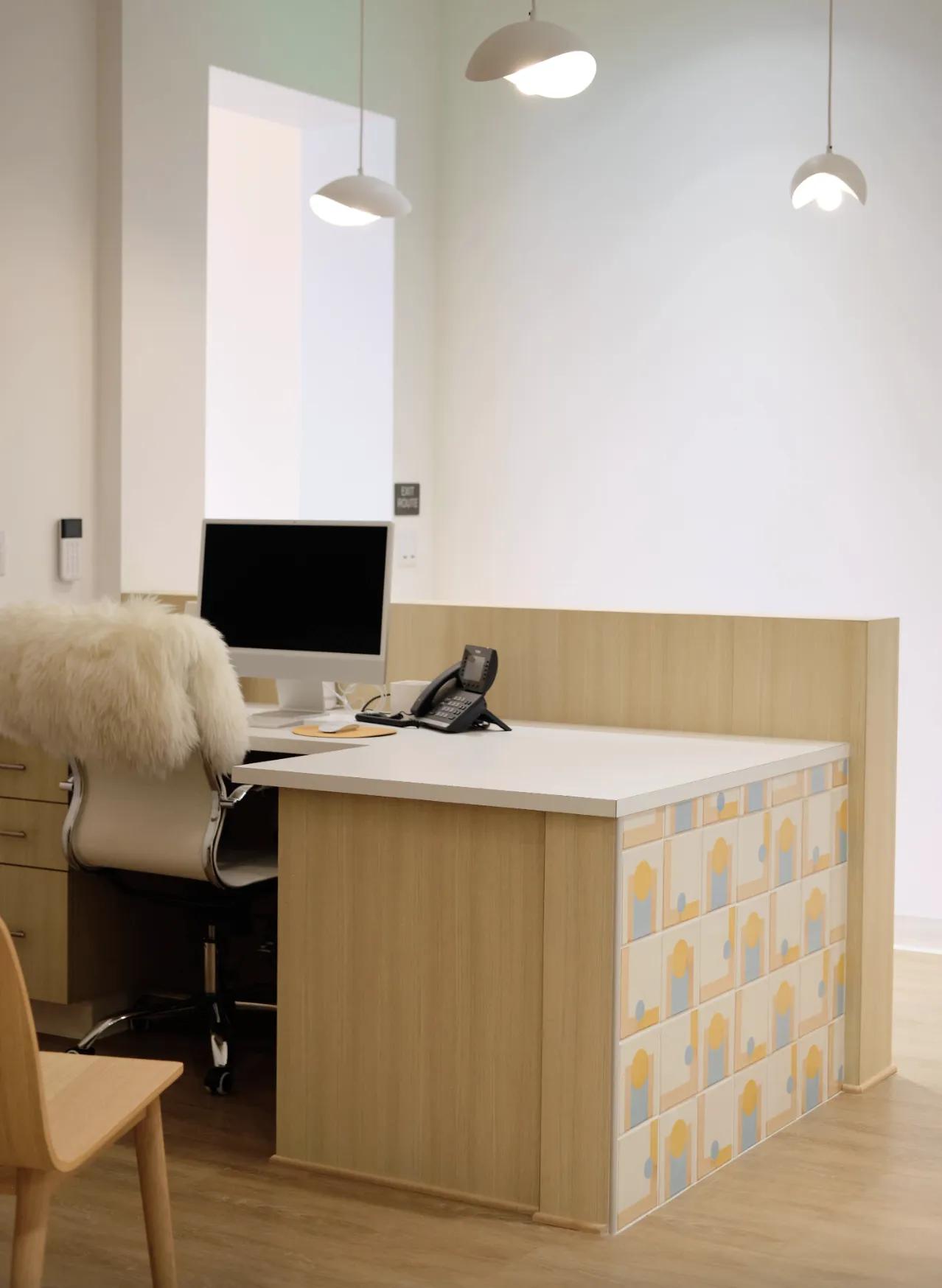Poppy Kids Dental reception area with a modern, minimalist design. The desk is made of light wood with a white countertop decorated with a colorful geometric pattern. A computer monitor, phone, and a cozy white chair with a fur throw are situated at the desk. Above, white pendant lights illuminate the space, and a clean white wall provides a calm background.