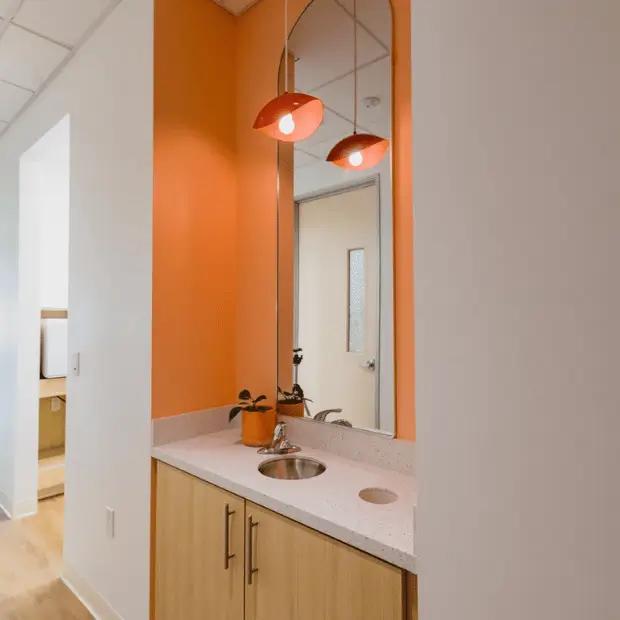 An image of Poppy Kids Dental treatment hallway with a light wood cabinet and a speckled countertop, which includes a built-in circular sink. Above the sink, a trio of red pendant lights adds a warm glow to the orange and white walls. A tall mirror extends from the countertop to the ceiling, reflecting the interior space. The entrance to another room is visible on the left, and the floor is covered in light wood laminate.