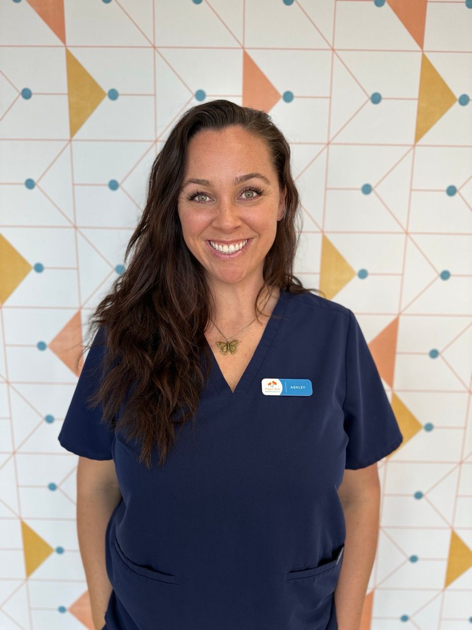 Poppy Kids office coordinator Ashley sits smiling on a large, weathered tree trunk in a sunlit field with green grass and trees in the background.