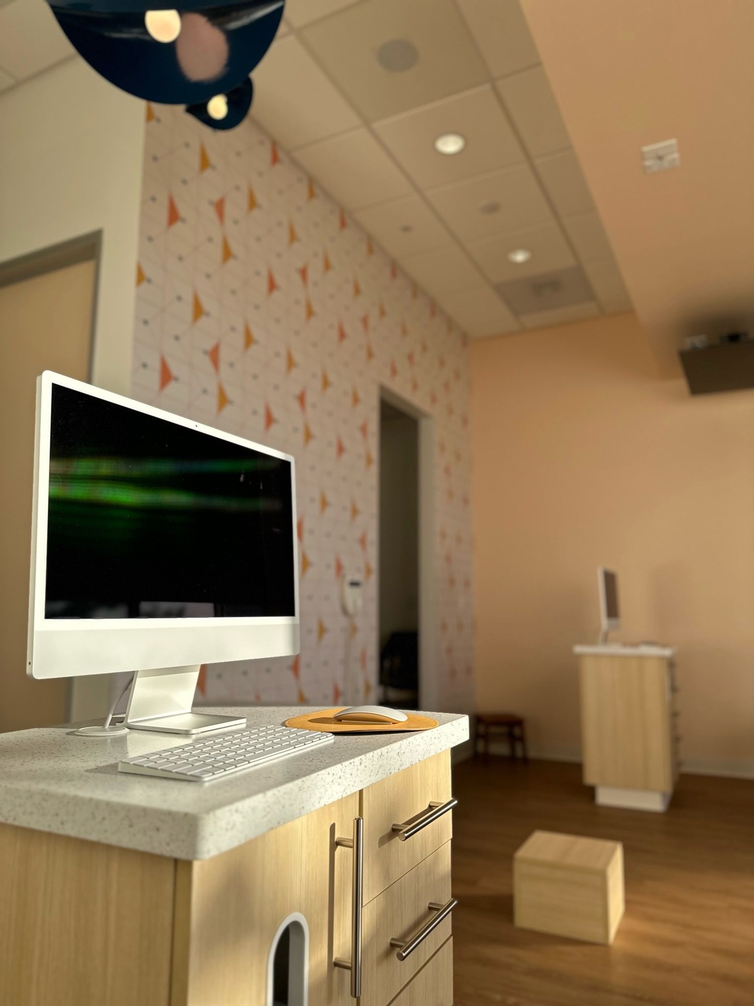A warmly lit dental office workstation with a wooden desk and a modern computer monitor. In the background, a wall adorned with playful kite-patterned wallpaper in orange and gray hues adds a cheerful atmosphere. The floor is a warm wood tone, and the room is well-lit with recessed ceiling lights. A second desk and computer setup can be seen in the adjoining room through the open door.