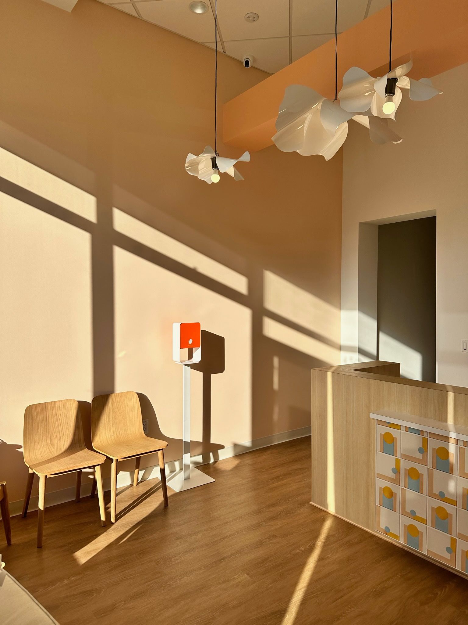 The interior of a dental clinic reception area bathed in warm sunlight with long shadows cast across the floor. There are two simple wooden chairs and a temperature check station with a red sign. Above, white petal-shaped pendant lights hang from the ceiling, and a wooden reception desk with a colorful front panel completes the welcoming space.