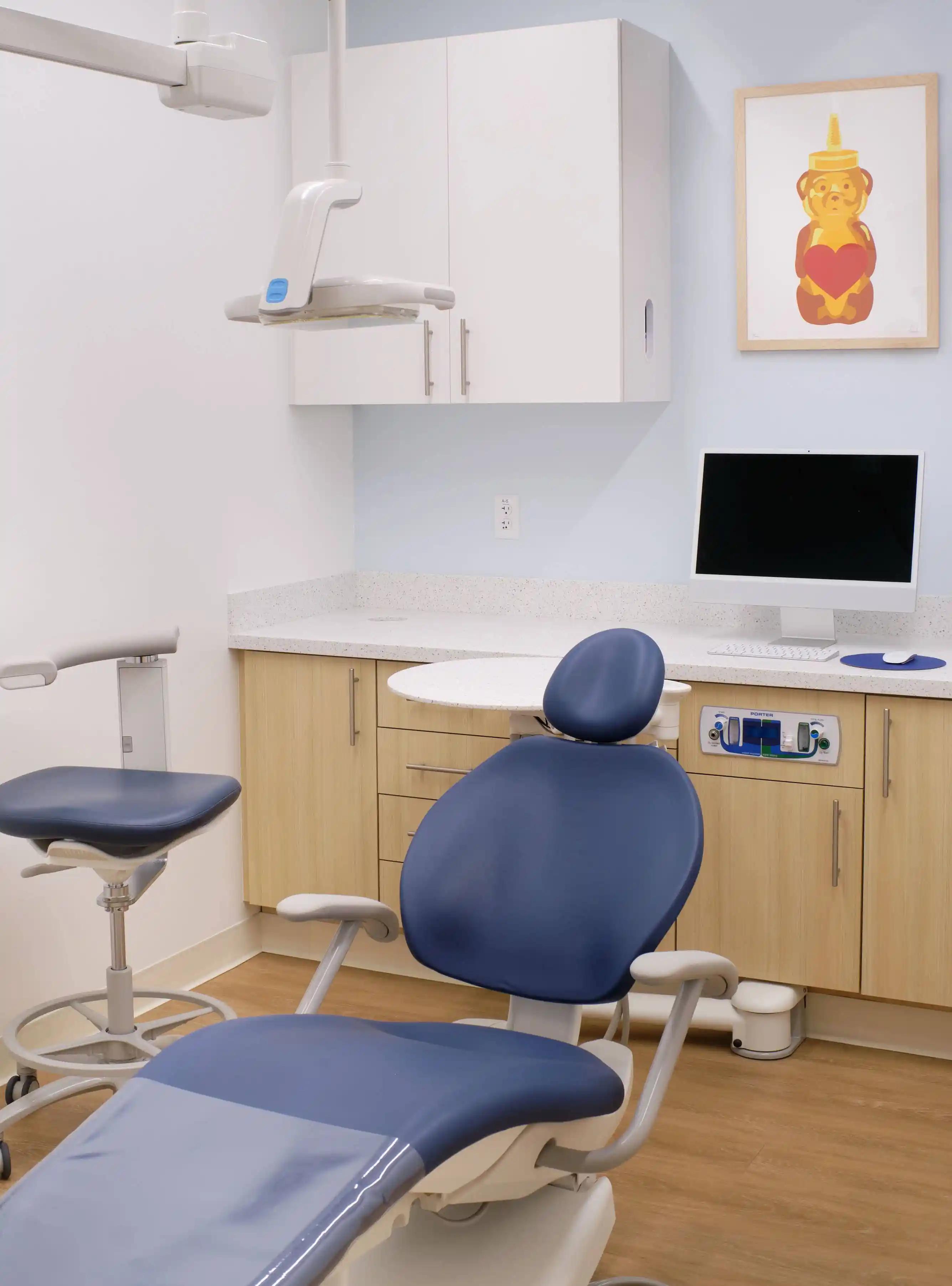 Poppy Kids Dental treatment room with modern equipment. A dental chair upholstered in blue is positioned in the center, with a matching practitioner's stool beside it. Above the chair, an adjustable dental light is mounted. The room features a curved white countertop with wooden cabinetry, a computer monitor, and built-in dispensers for gloves and tissues. On the wall, a framed picture of a cartoon bear with a heart by San Francisco artist fnnch adds a child-friendly touch to the space.