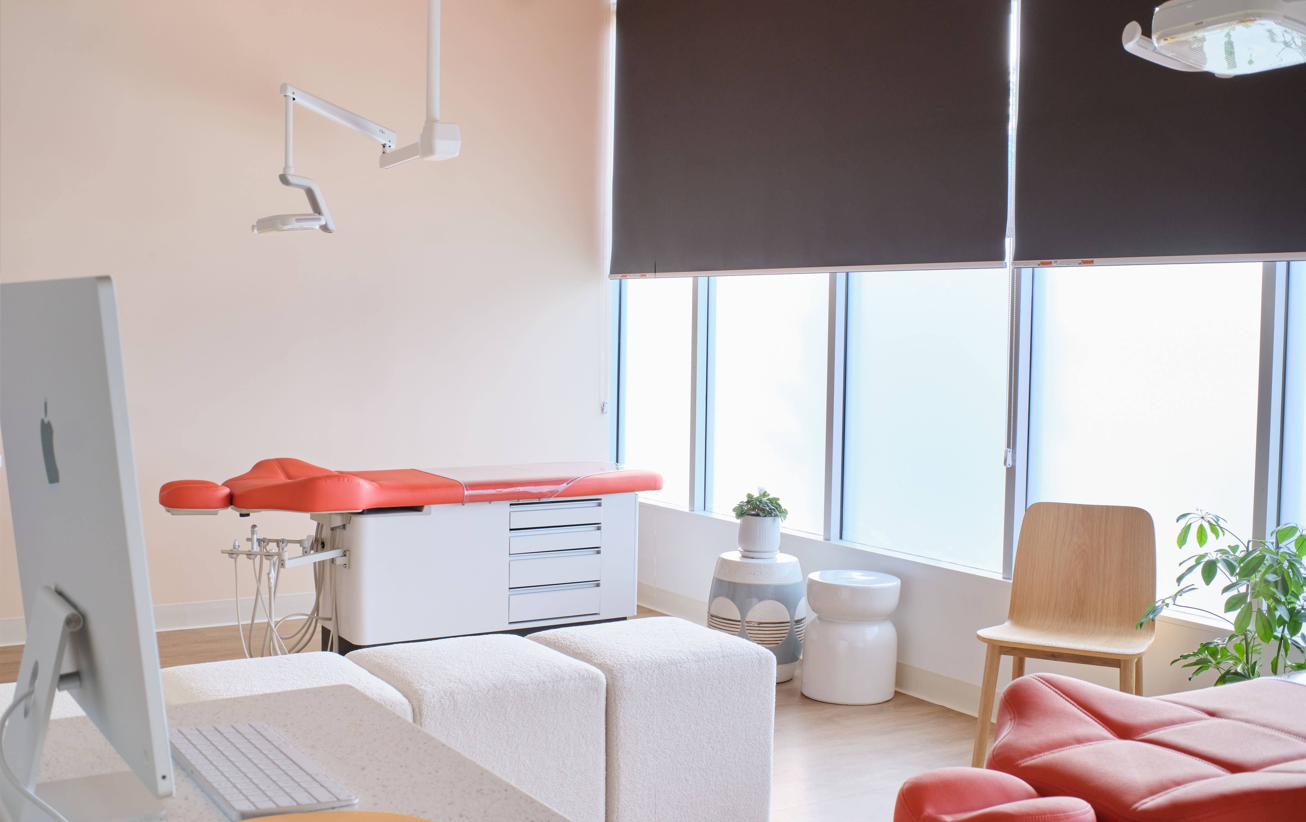 A bright, modern dental office with large windows covered by partially drawn brown blinds. The room features a dental chair with orange upholstery, a white dentist's work station with a computer monitor, a simple wooden chair, and a white waiting bench. Decorative elements include a small potted plant on a patterned vase and a larger green plant, creating a comfortable and inviting atmosphere.
