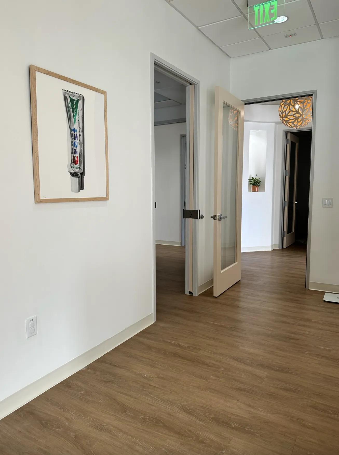 A view of a dental clinic's hallway featuring wood-like flooring and white walls. On the left wall, there's a framed artwork of a large toothpaste tube. Glass doors open to various rooms within the clinic, and a unique spherical orange light fixture hangs in the corridor. A green exit sign is visible at the ceiling, indicating the way out.