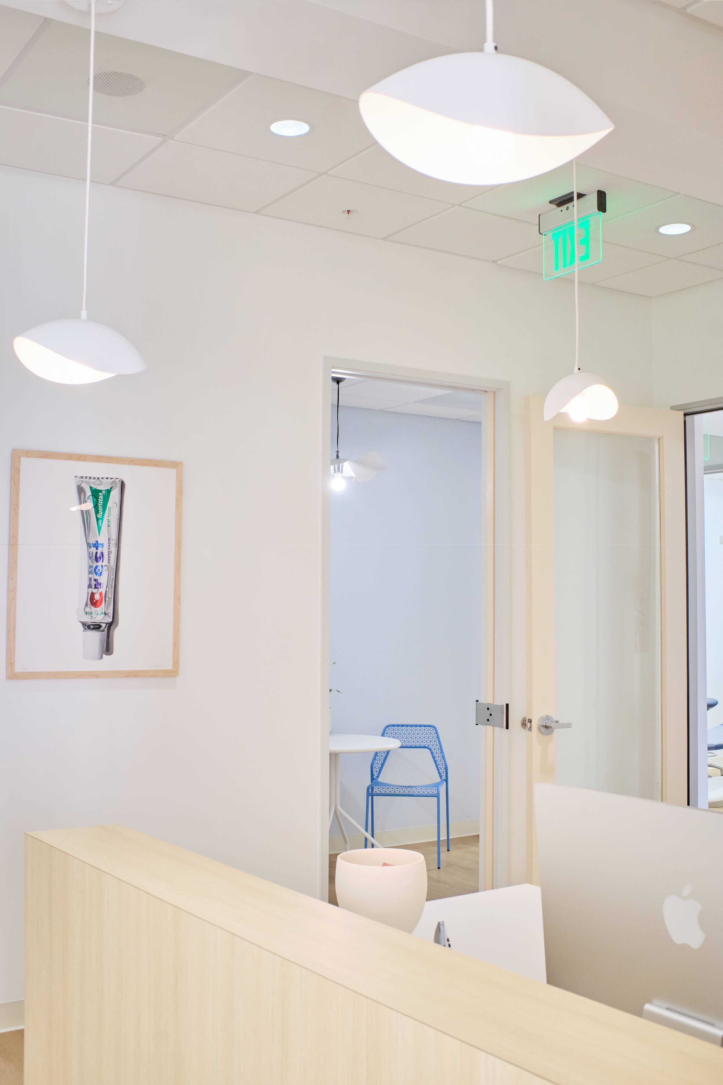 A bright and airy dental office reception area. The room features white walls, modern hanging white pendant lights, and a light wood reception desk with an Apple computer on it. On the left wall, there's a framed artwork resembling an oversized tube of toothpaste. A blue patterned chair sits in the adjacent room visible through the open door, and a green exit sign glows above the doorway.