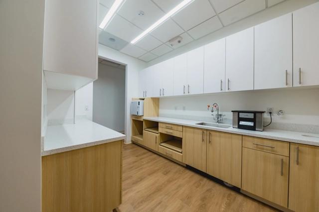 The image displays a modern sanitary room at Poppy Kids Pediatric Dentistry. The room features clean, well-lit spaces with white and light wood cabinetry. It includes multiple sinks with ample countertop space, a sterilization machine, and lighting that highlights the pristine condition of the facility.