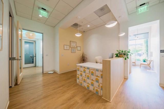 The hallway of Poppy Kids Dental with warm wooden floors and a reception desk to the right with a toothbrush and cup pattern. The desk is accompanied by a plant with broad green leaves. The wall displays framed certificates, and the area is well-lit with dome-shaped pendant lights and a green exit sign at the end of the corridor. An open door on the left gives a glimpse into another room, and a waiting area with wooden chairs is visible through the doorway at the far end.