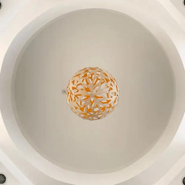 An intricate, spherical light fixture hangs from the center of a white, circular dome ceiling at the Poppy Kids Dental. The fixture has a patterned design with interlacing yellow and white elements that create a lattice-like appearance. The dome is edged with geometric cutouts and recessed lighting, giving a modern and elegant look to the architecture.