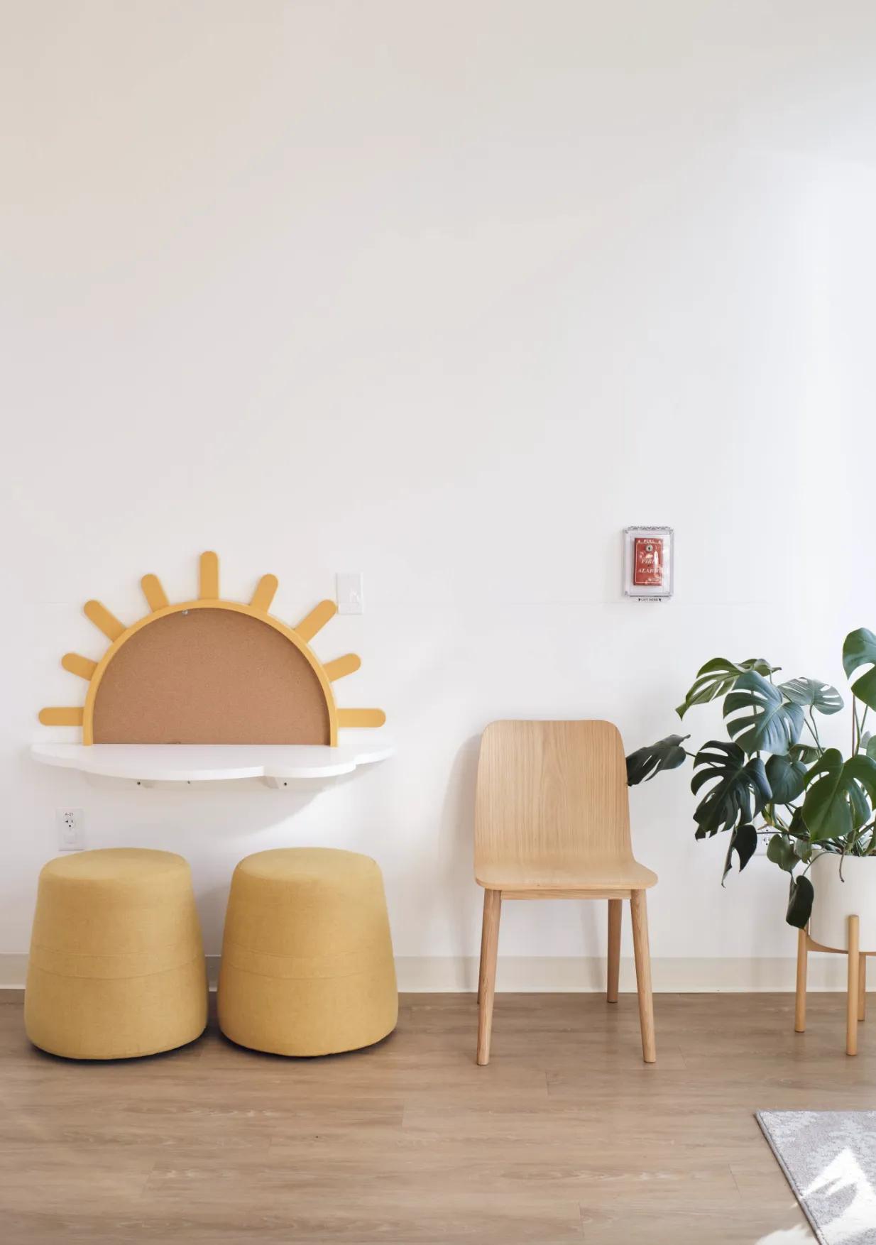 Poppy Kids Dental children's area with a playful sun-shaped bulletin board on the wall. Below it, two mustard yellow poufs provide seating next to a simple wooden chair. A lush green potted plant adds a touch of nature to the room, which has a clean and bright aesthetic with white walls and light wooden flooring. A red fire alarm pull station is mounted on the wall for safety.