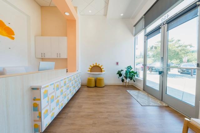 Poppy Kids Dental's sunny waiting area with a large windowed entrance door. To the left, a reception desk showcases a playful design with toothbrush and cup patterns. The wall features a floral artwork, and there is a child-friendly bench with a sun-shaped backrest and two round yellow stools. A healthy potted plant is situated near the entrance, enhancing the cheerful and welcoming atmosphere of the clinic.
