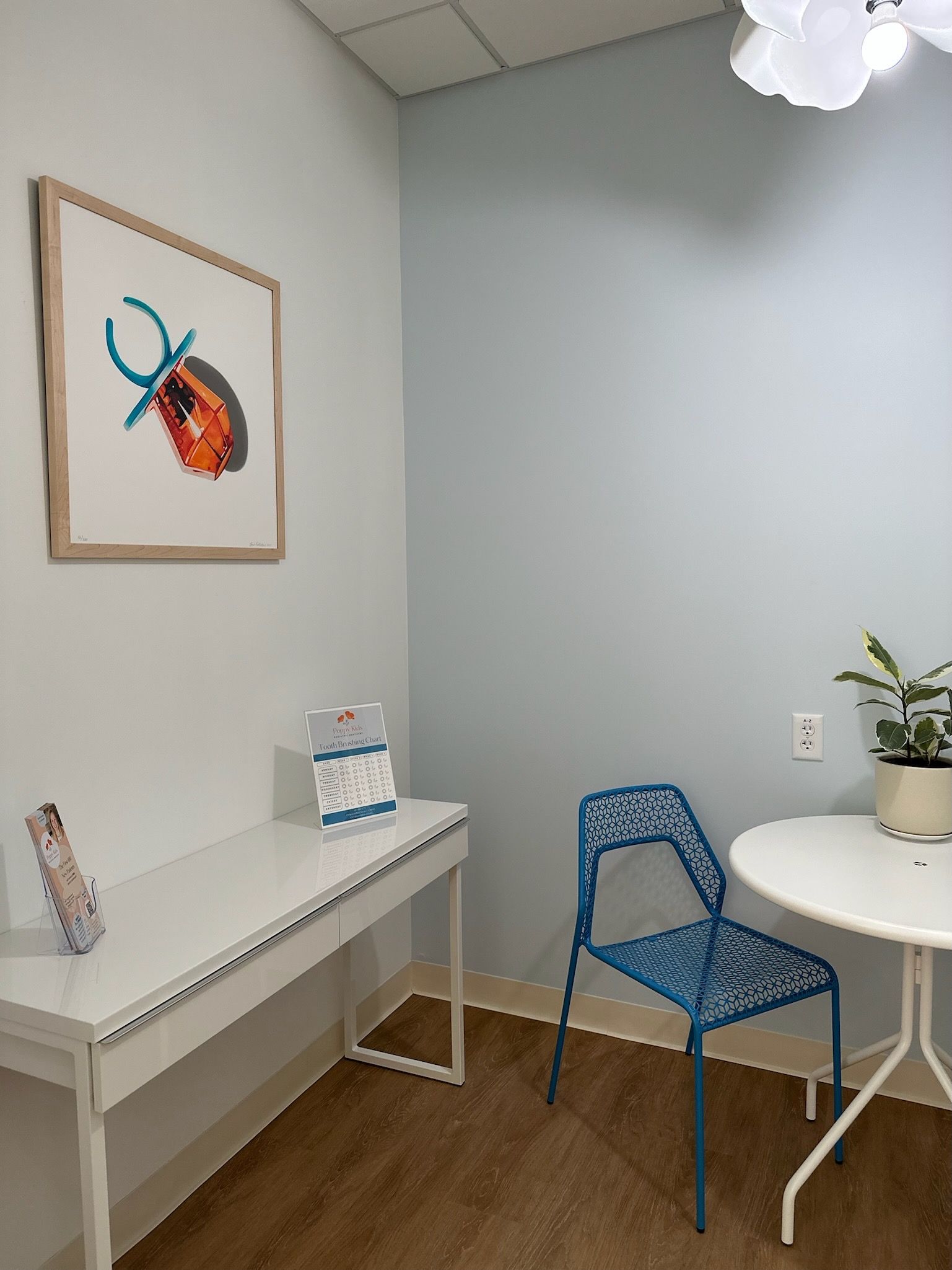 A consultation room with pale blue walls and wooden flooring. A white modern desk with information leaflets and a round table with one blue patterned chairs is placed in the room. On the wall, there's a framed artwork of a colorful pacifier made from candy, and above, a unique white floral pendant light fixture illuminates the space.