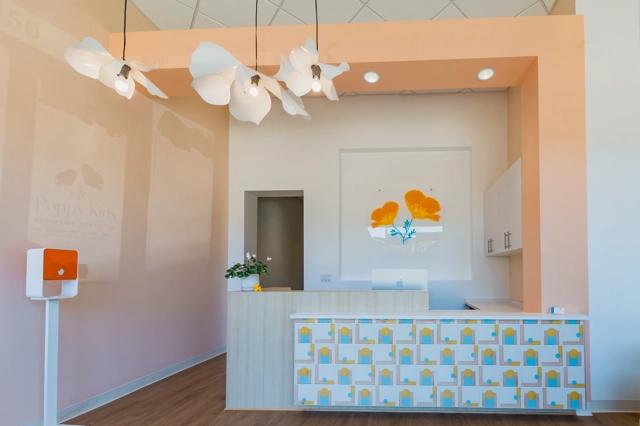Interior view of Poppy Kids dental clinic's reception area with a light wood front desk, featuring a colorful pattern of toothbrushes and cups. Above, unique flower-shaped lights hang from the ceiling. A graphic with 'Poppy Kids Pediatric Dentistry' is visible on the wall, and a digital check-in screen stands to the left. The back wall showcases an artistic floral design visible through a large cut-out window.
