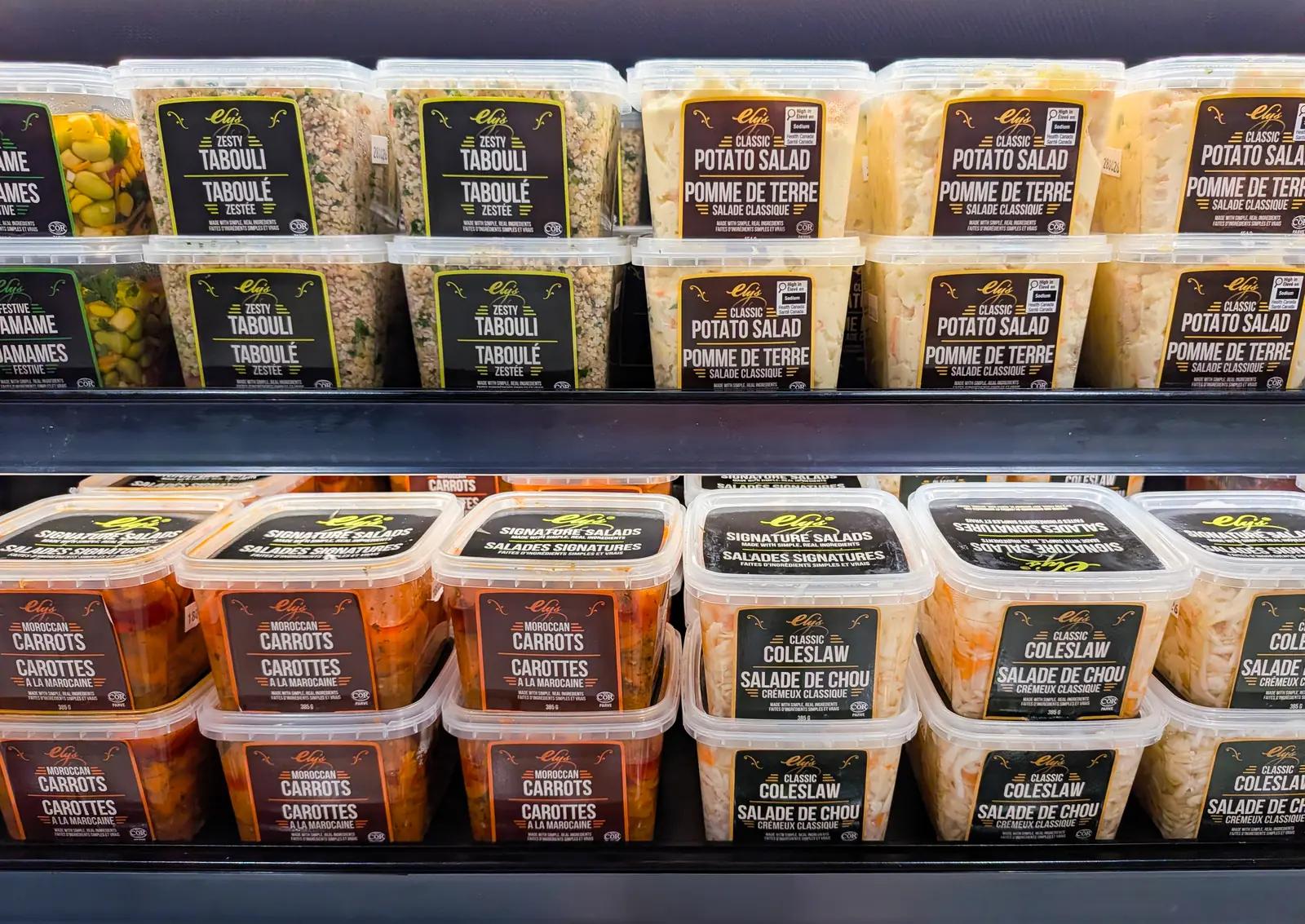 Cooler shelf of Ely's salads in the new labels: Zesty Tabouli in black and lime, Classic Potato Salad in brown and gold, Moroccan Carrots in maroon and orange, and Classic Coleslaw in dark green and gold
