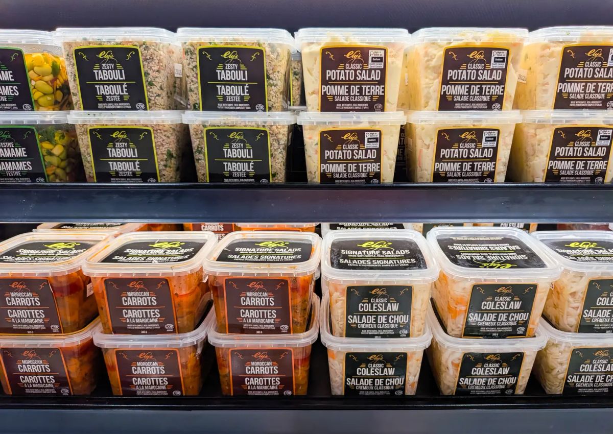 Cooler shelf of Ely's salads in the new labels: Zesty Tabouli in black and lime, Classic Potato Salad in brown and gold, Moroccan Carrots in maroon and orange, and Classic Coleslaw in dark green and gold