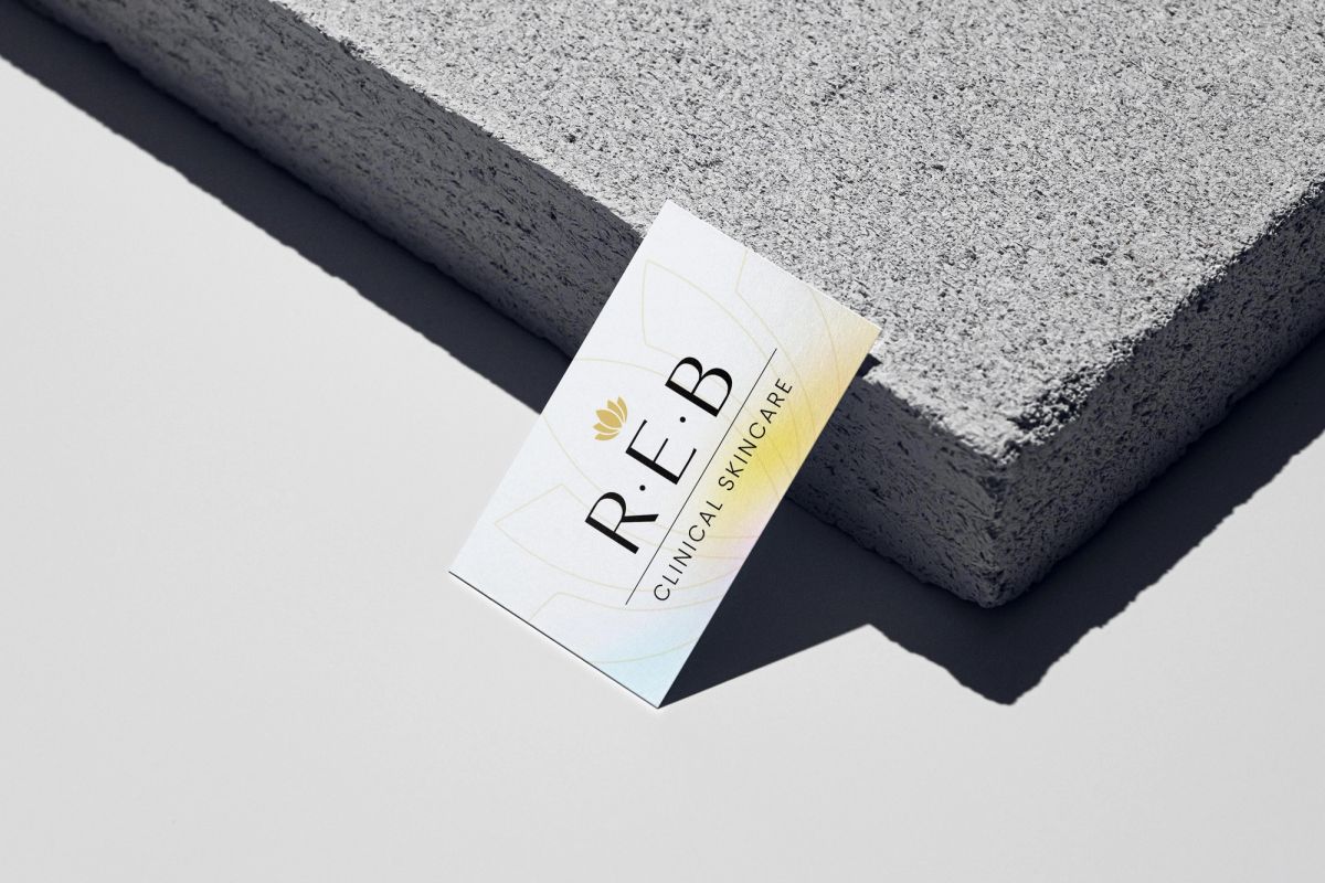 REB business card with a soft iridescent edge, leaning on grey concrete