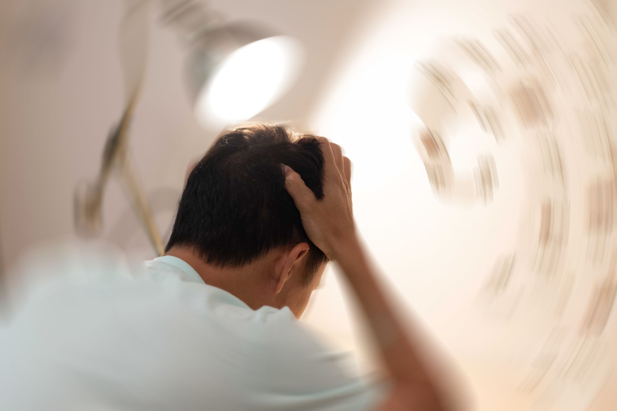 Mann med kort brunt hår og hvit t-skjorte sitter med ryggen mot kamera og griper seg til hodet, bakgrunnen er uklar og i sirkelbevegelse rund mannens hode.