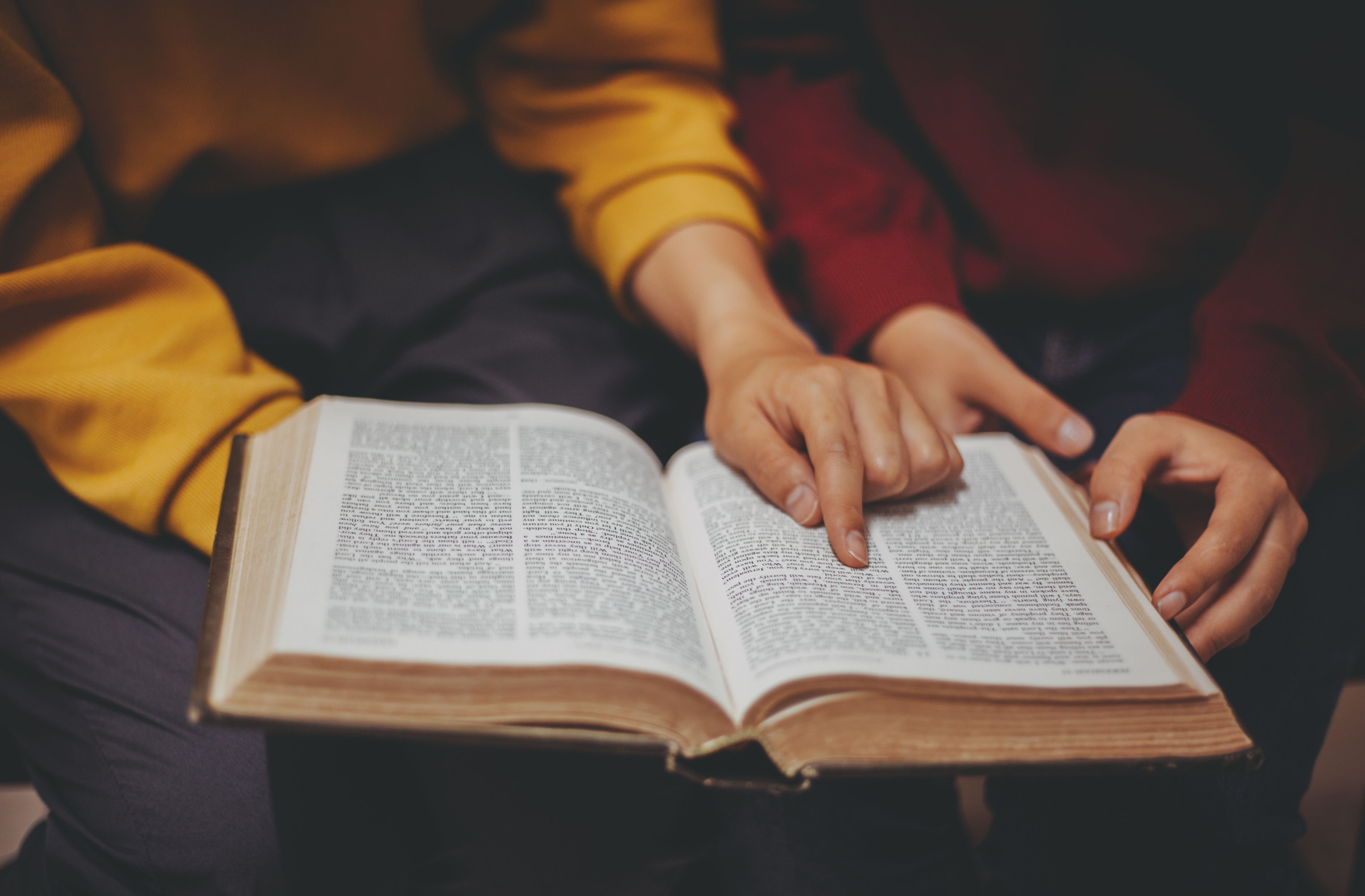 Foto, en åpen bibel. To personer leser i den sammen. De holder boken over knærne mellom seg og peker på ett ord på siden.