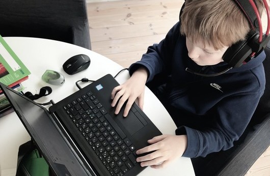 Foto, en gutt i barneskolealder som sitter med en laptop og har hodetelefoner på. Sett ovenfra.