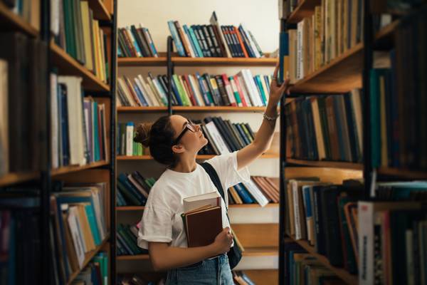 En student som holder pensumbøker i hånden og leter etter bøker på biblioteket.