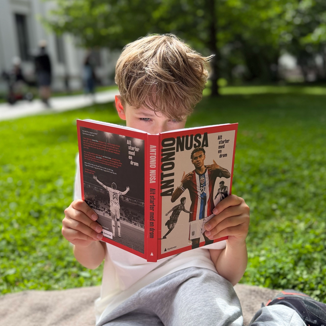 Gutt som sitter på et pledd ute og leser i en bok