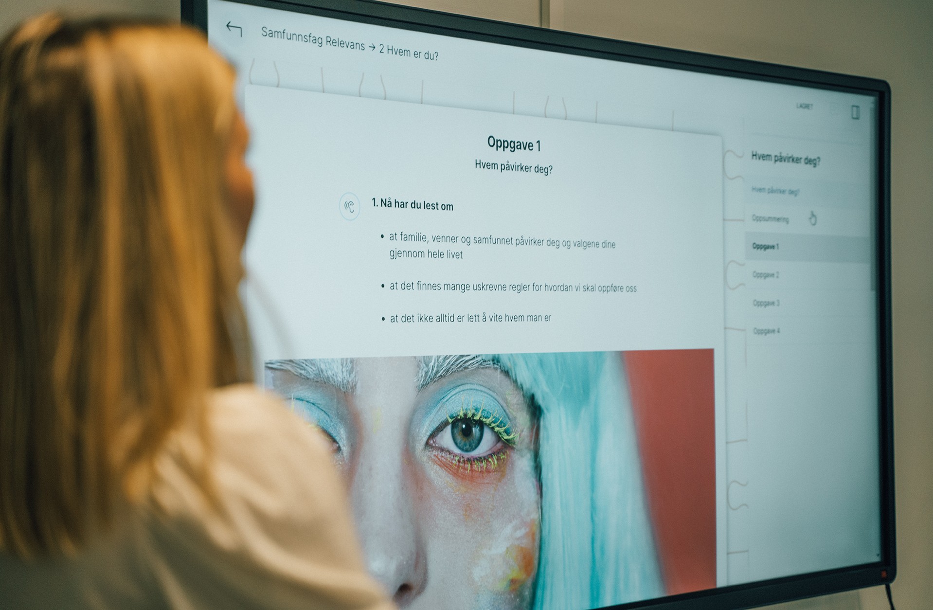 Smartboard tavle med en side fra Skolestudio på skjermen. En kvinne med lyst hår står til venstre i bildet og ser på skjermen. Hun blir sett bakfra, og er ute av fokus.