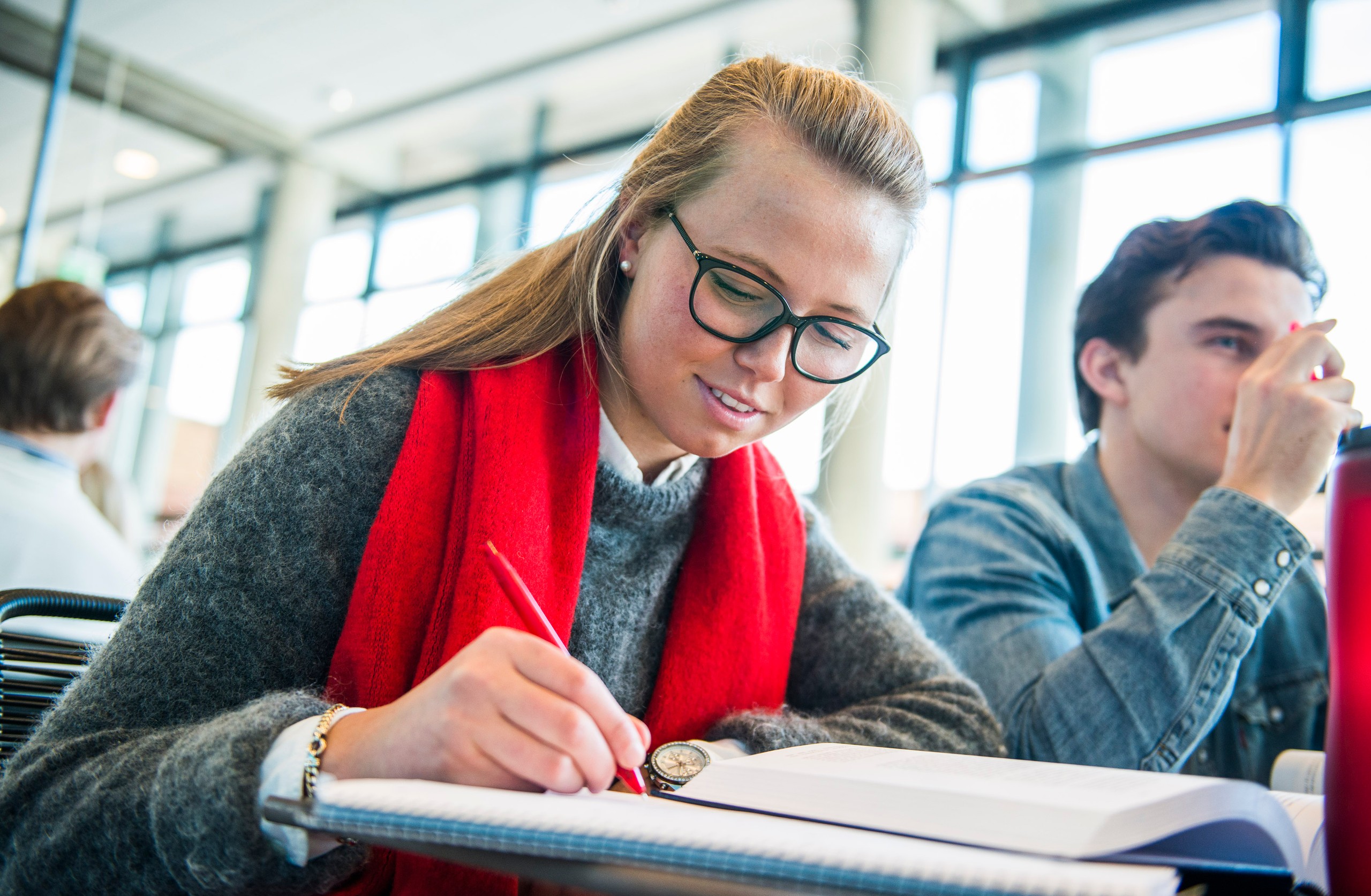 Illustrasjonsfoto. Studenter. I forgrunnen er en kvinnelig student, med en bok åpen på bordet foran seg, som skriver i en notatblokk.