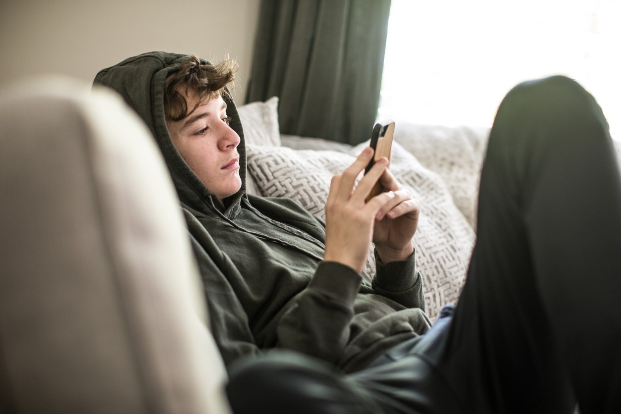 Tenåringsgutt i grønn hettegenser og olabukse sitter i en sofa og bruker en mobiltelefon.
