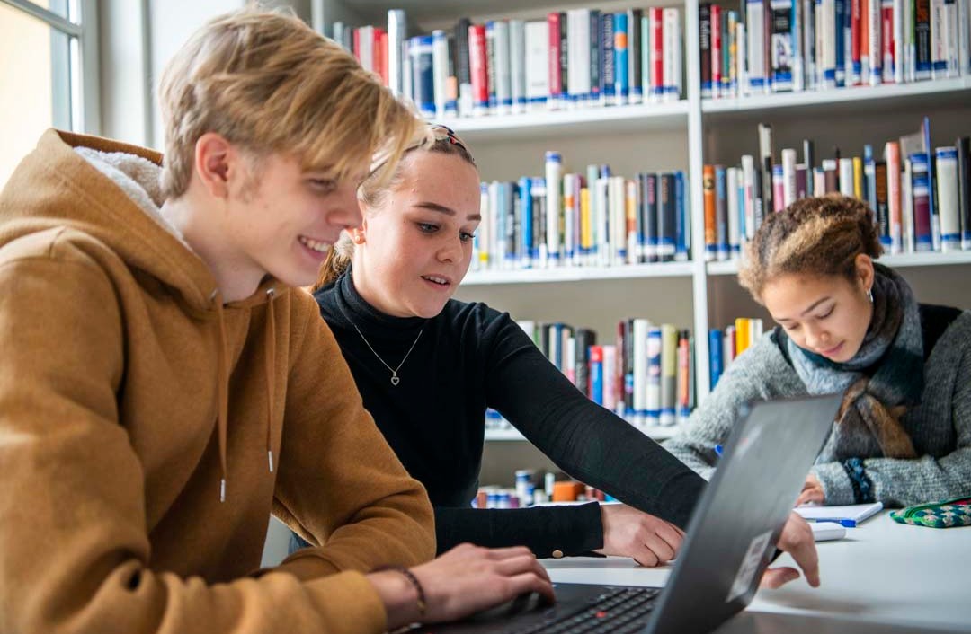 Foto, tre unge voksne som sitter ved ett bord og jobber. To fokuserer på en laptop, mens den siste sitter med en skriveblokk.