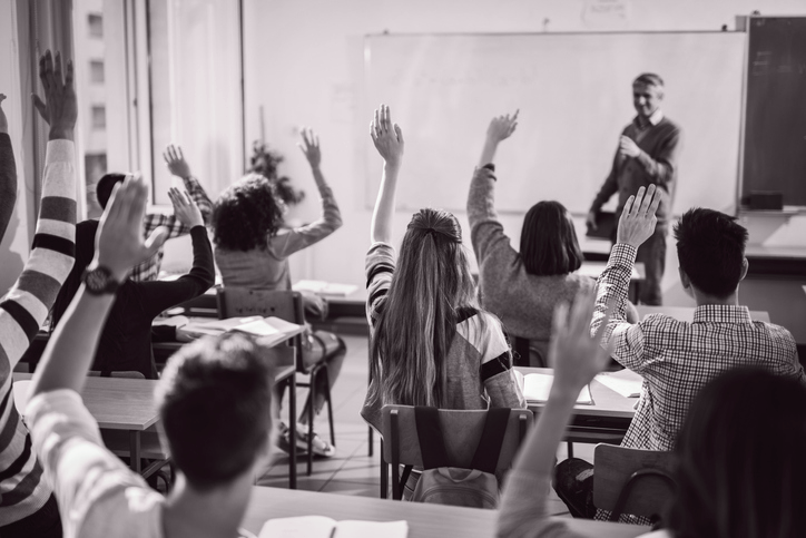 Svart-hvitt bilde av ett klasserom med ungdommer som rekker opp hånden og en lærer som står foran klassen. Bildet er tatt fra bakveggen i klasserommet og læreren er ute av fokus.