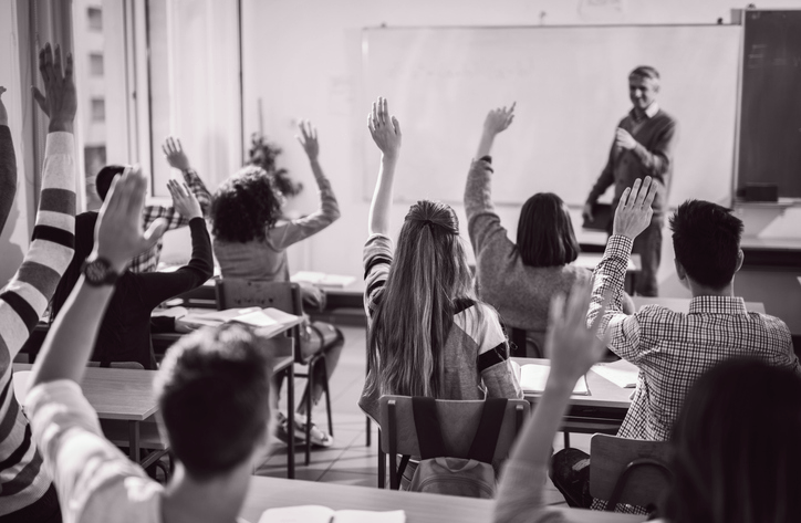 Svart-hvitt bilde av ett klasserom med ungdommer som rekker opp hånden og en lærer som står foran klassen. Bildet er tatt fra bakveggen i klasserommet og læreren er ute av fokus.