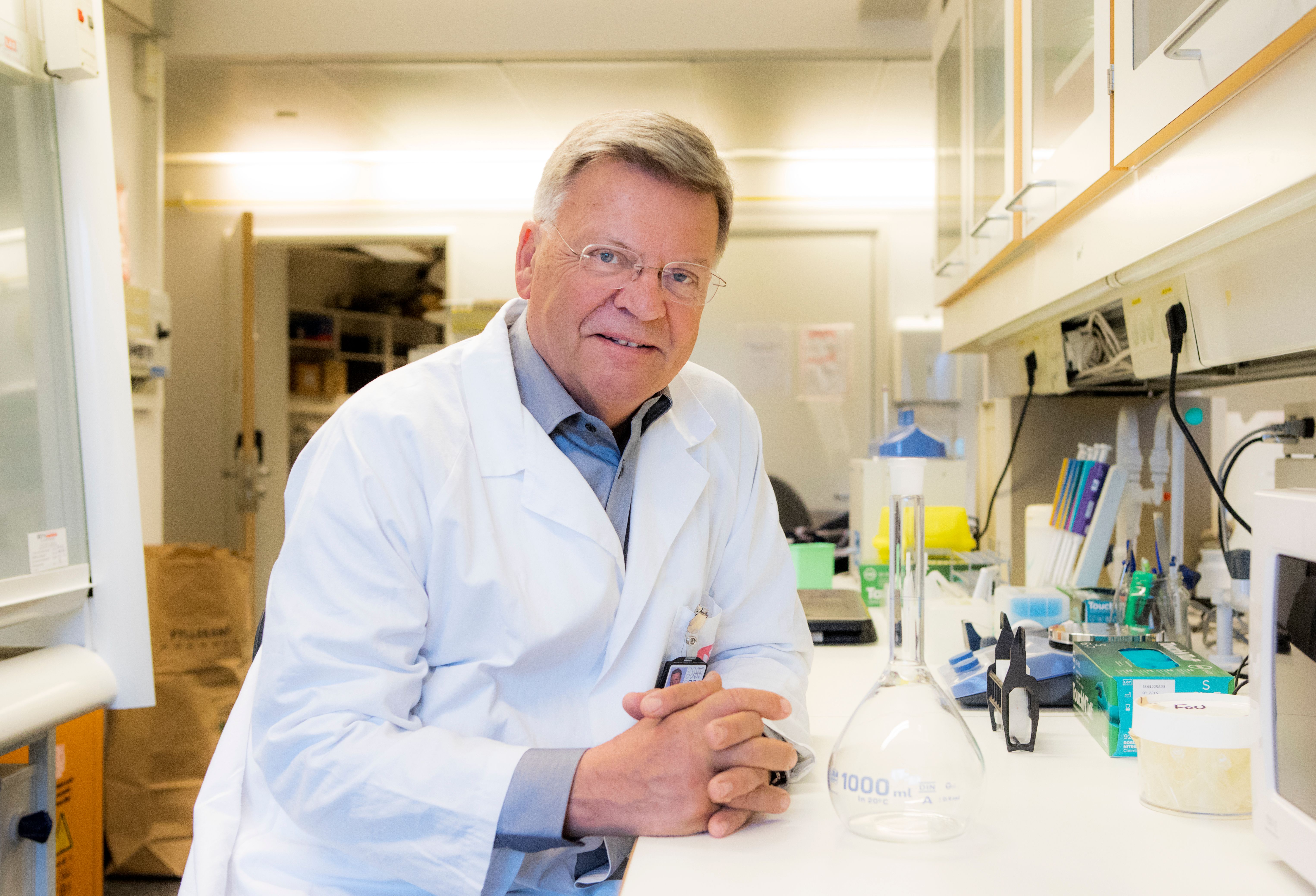 Foto av Torleiv Rognum. Han har labfrakk på seg, og sitter vendt mot kamera mens han lener seg på en laboratoriebenk.