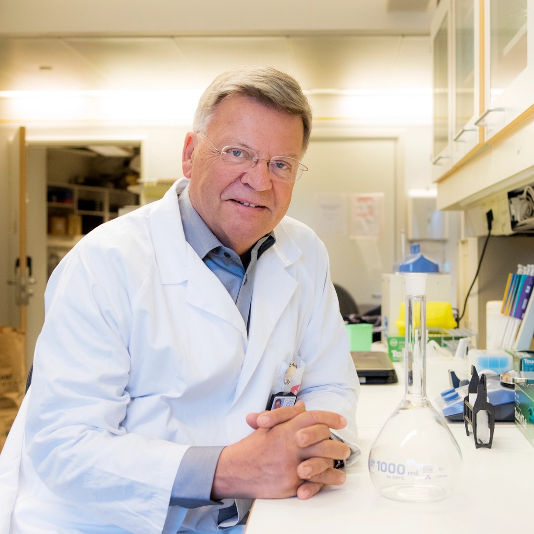 Foto av Torleiv Rognum. Han har labfrakk på seg, og sitter vendt mot kamera mens han lener seg på en laboratoriebenk.