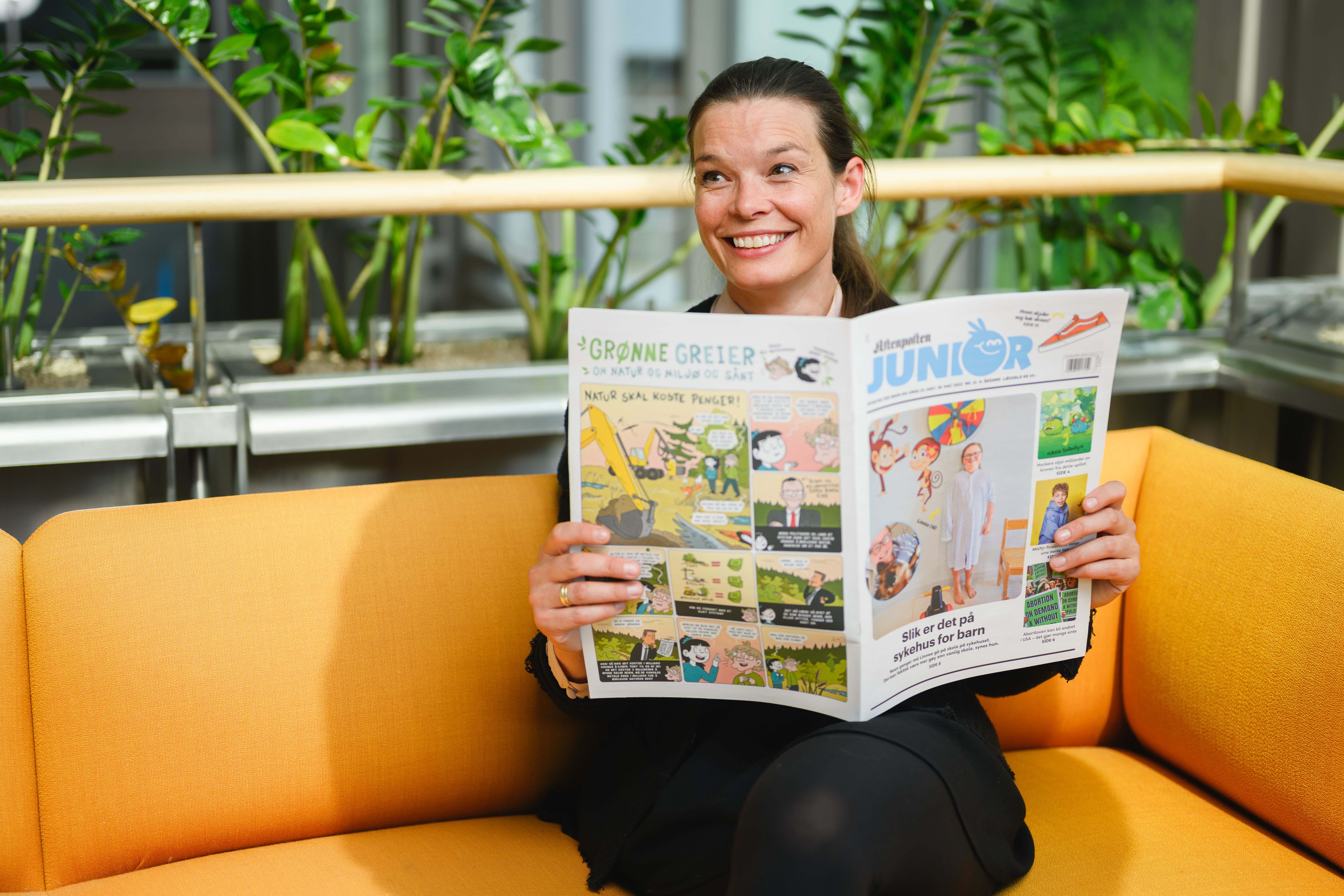 Mari Midtstigen, redaktør i Aftenposten Junior Skole, sitter på en sofa og holder opp ett eksemplar av Aftenposten Junior.