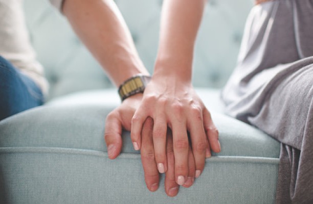 To personer sitter tett sammen i en sofa og holder hender. Bildet illustrerer nærhet, støtte og samspill i et parforhold der én har ADHD.