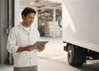 A driver holding a tablet beside a cargo truck at a sunlit loading bay.