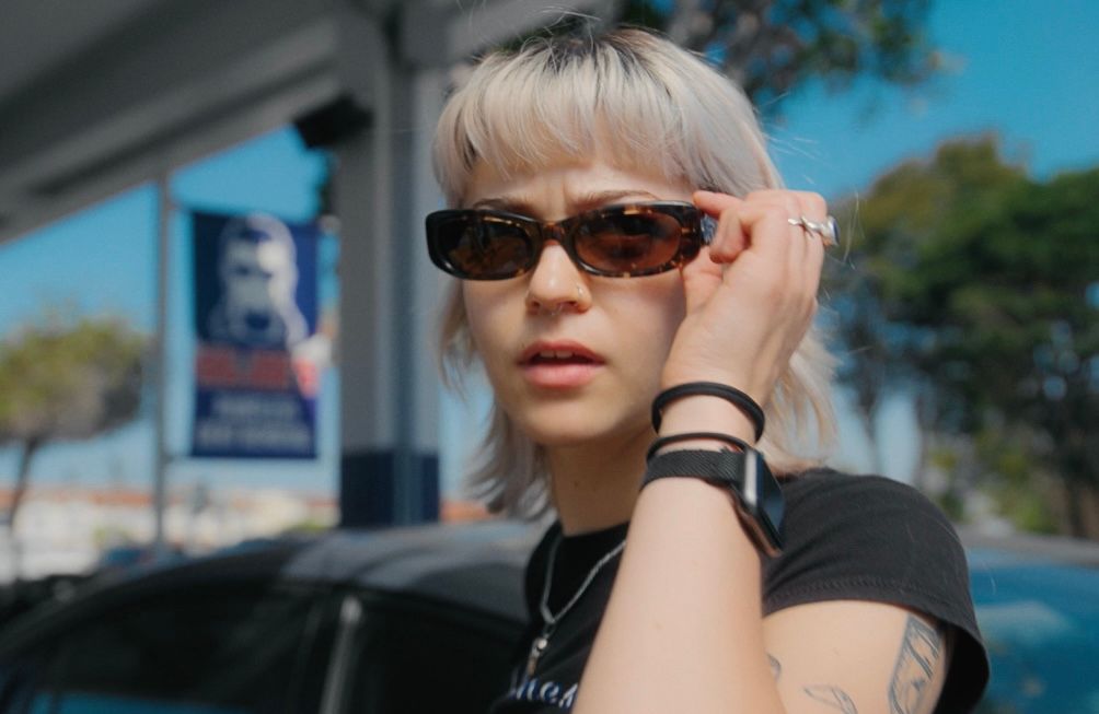 Emmy puts her hand up to her sunglasses, pausing in anticipation.