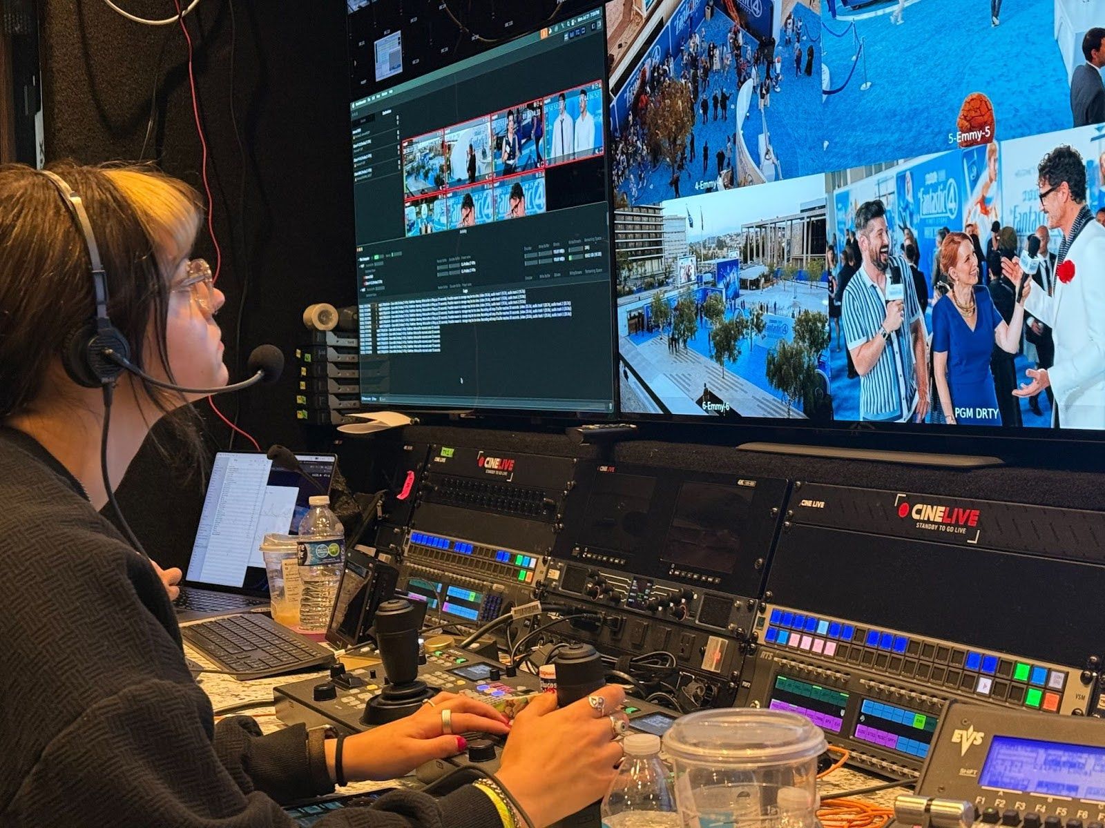 Emmy sits at a desk with both hands on the camera operation controls while looking at a screen displaying four camera perspectives, one including a shot of Pedro Pascal.