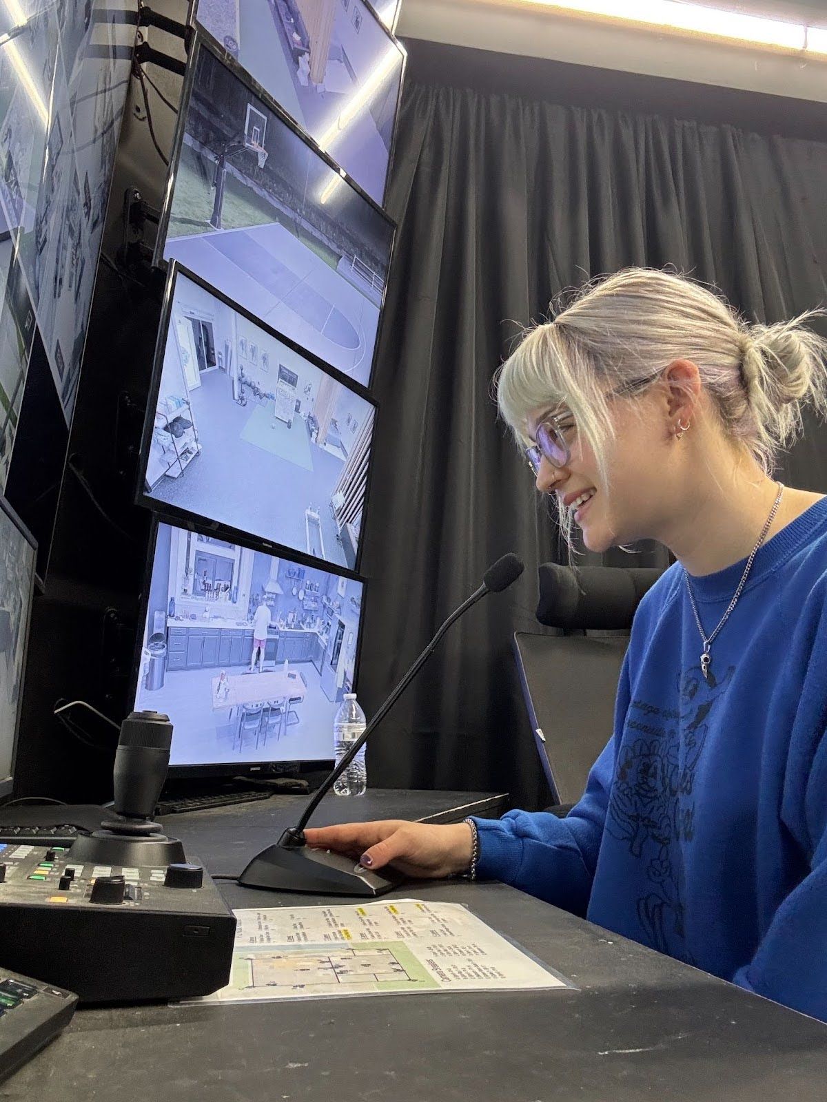 Emmy sits at a desk in a camera operation control room, ready to speak into a microphone.