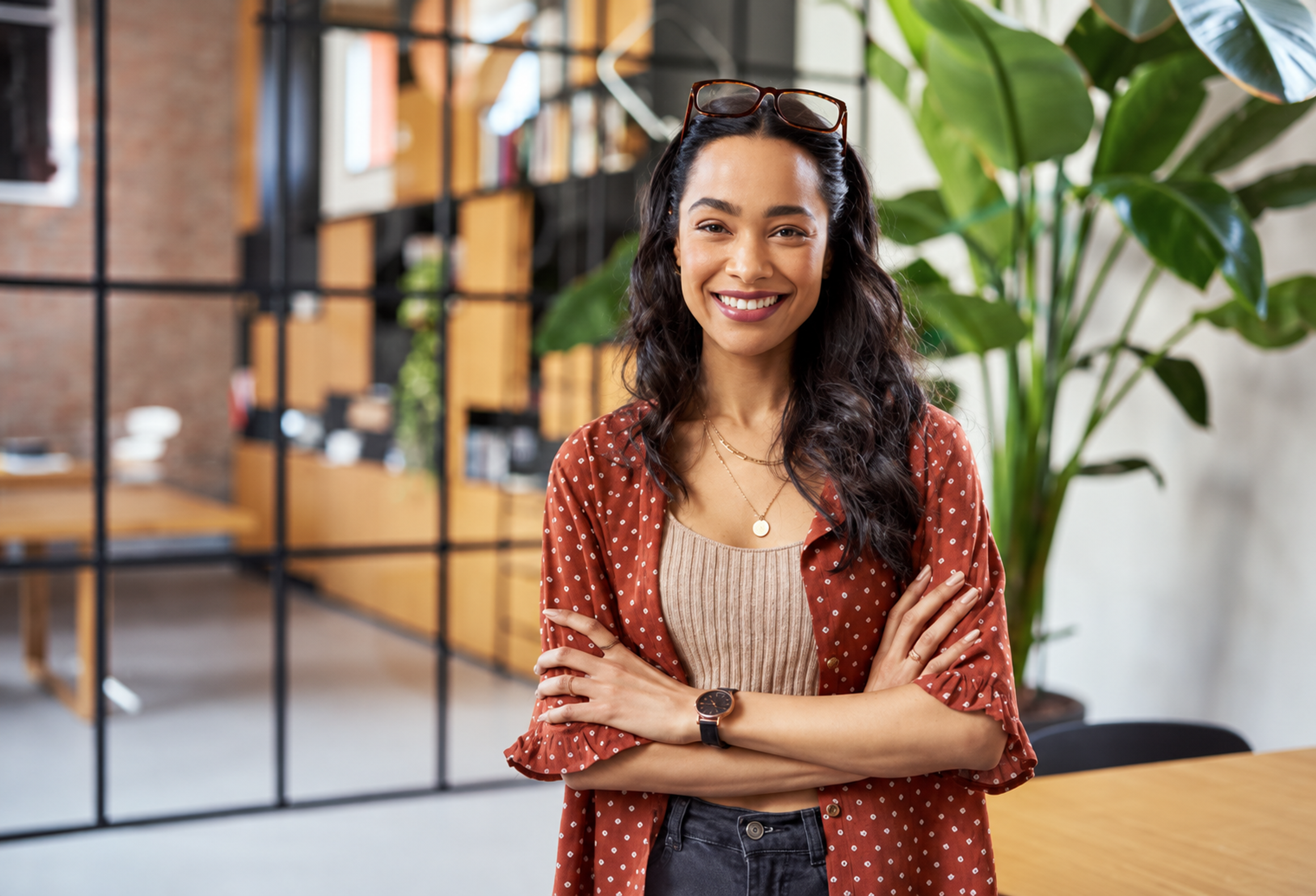 A smiling woman with glasses on her head and arms crossed in a modern office.