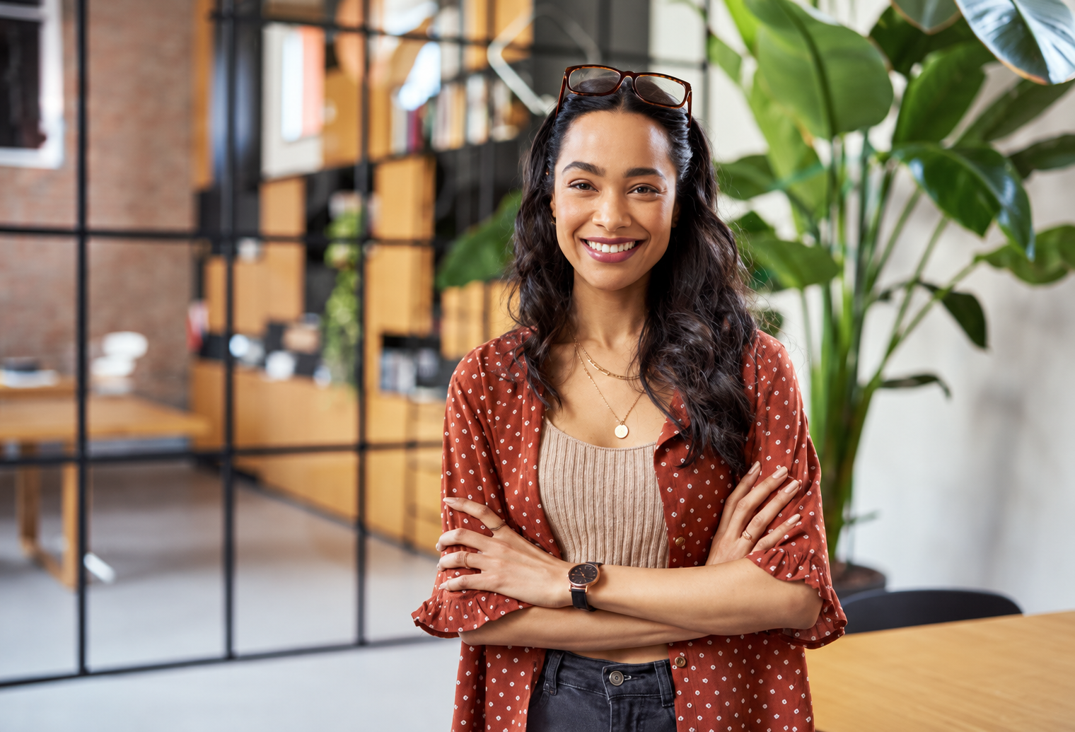 A smiling woman with glasses on her head and arms crossed in a modern office.