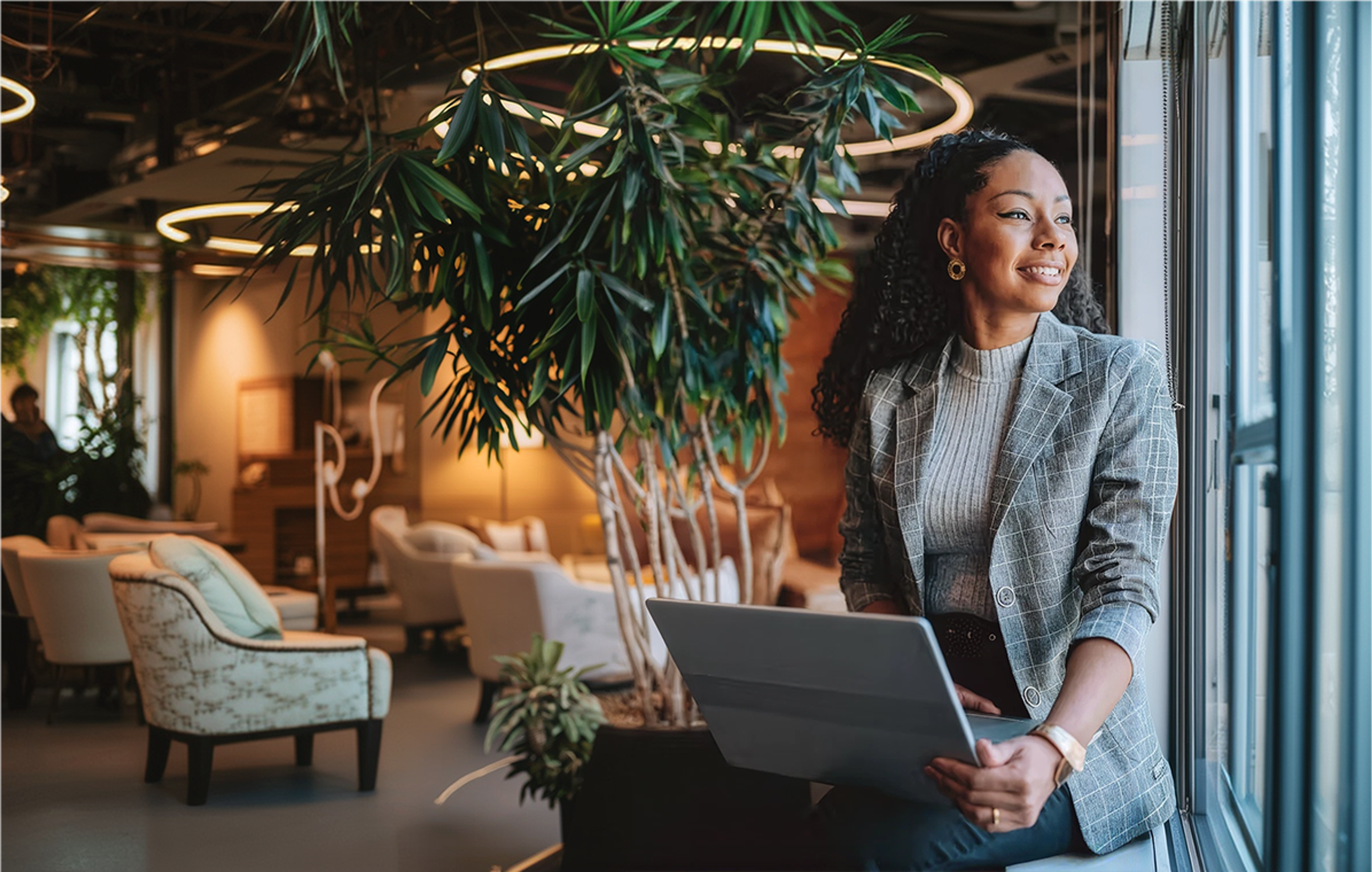 A professional woman in a gray plaid blazer smiling and holding an open laptop near a window next to a houseplant.